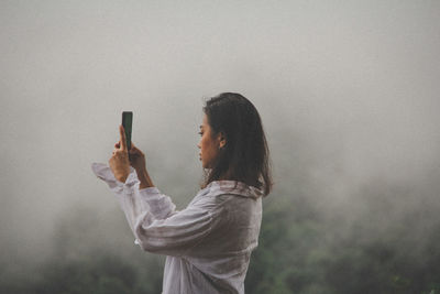 Side view of young woman photographing from mobile phone during foggy weather