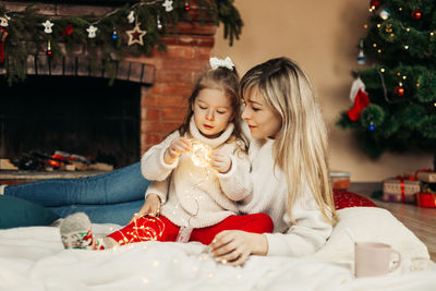 Mom and daughter are playing with christmas garlands on the floor by the fireplace