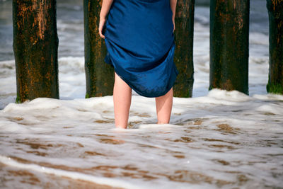 Legs of young beautiful woman in long dark blue dress standing in water on sandy beach, close up