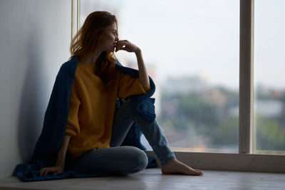 Young woman using phone while sitting on window at home