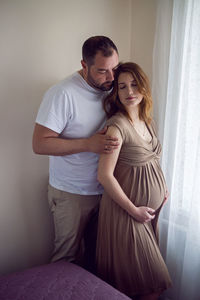 Pregnant girl in a brown dress is standing at the window with her husband in a white t-shirt at home