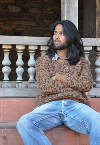 A long haired young guy posing with crossed arms, looking sideways while sitting on temple stairs 