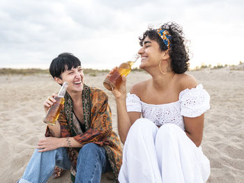 Positive multiethnic female best friends drinking cold beer while sitting on sandy seashore and laughing in summer evening