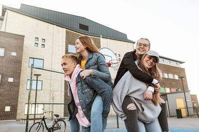 Smiling teenage boy and girl piggybacking friends while standing against building in city