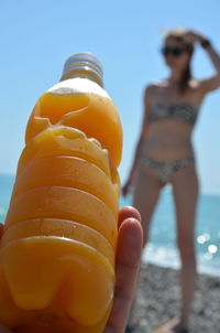 Cropped hand holding bottle while woman in bikini standing in background