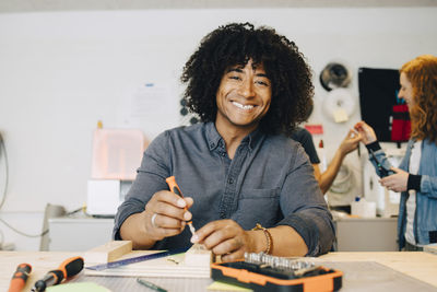 Portrait of smiling male technician using tool on wood at workbench in creative office