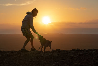 Woman playing with dog against sky during sunset