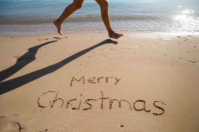 Low section of person running by merry christmas text at beach on sunny day