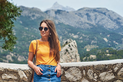 Young woman wearing sunglasses on retaining wall against mountains