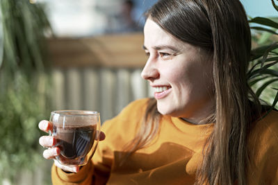 Smiling young caucasian woman holding glass cup of coffee on background of green plants, hot drinks