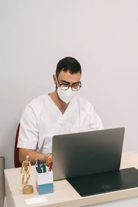 High angle of busy male physician in medical mask sitting at table and working on laptop in modern clinic