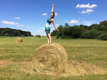 Cheerful woman balancing on hay bale at farm against sky