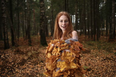 Portrait of smiling young woman covered with leaves standing at forest during autumn