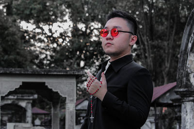 Young man wearing sunglasses holding rosary while standing against trees