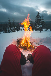 Low section of man sitting on snow by bonfire 