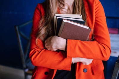 A young woman holds a stack of books. a girl in a red fashionable jacket against a blue wall.