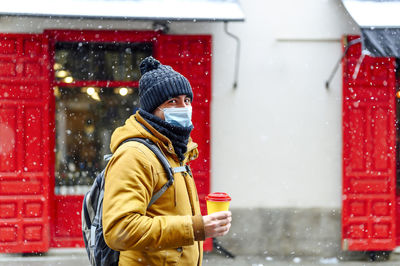 Man with coffee cup standing against store while snowing during pandemic