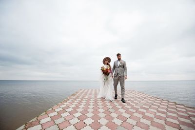 Couple walking on pier over lake against cloudy sky