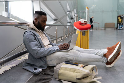 Young black man with laptop sitting in airport with suitcase searching information online about trip
