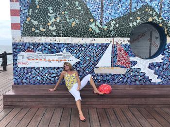 Full length of senior woman sitting against tiled wall on promenade