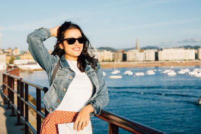 Portrait of smiling young woman standing by railing against sky