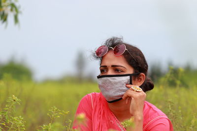 Young woman wearing mask looking away while sitting on field against sky