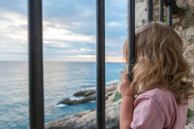 Girl looking through window at sea against sky