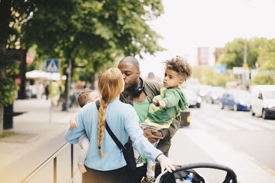 Parents kissing while carrying children on sidewalk in city