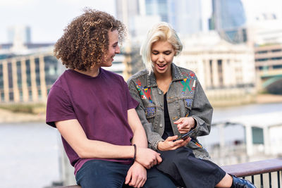 Woman using smart phone by friend while sitting on railing in city