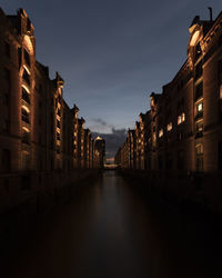 Illuminated street amidst buildings against sky at night