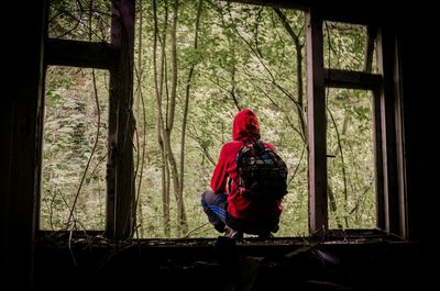 Rear view of hiker crouching on window of abandoned house by forest