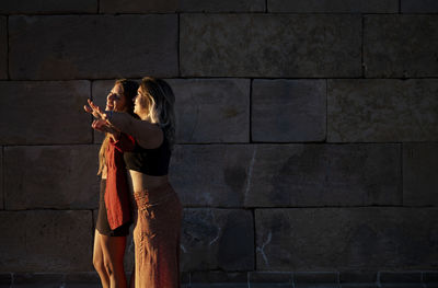 Two girls standing against stone wall during sunset