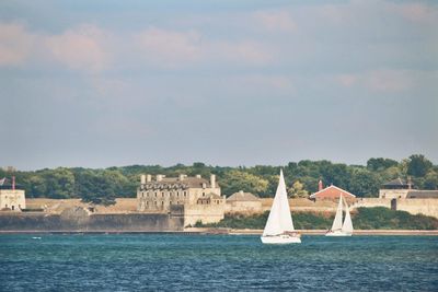 Sailboats sailing on sea by buildings against sky