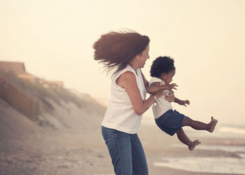 Side view of mother swinging daughter while standing at beach against sky