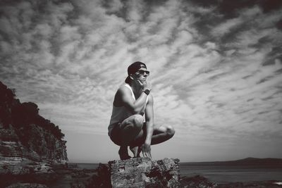 Young man crouching at beach against sky