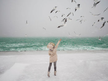 Woman reaching birds at beach against sky