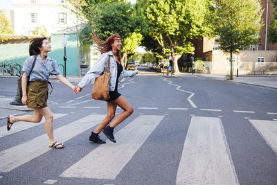 Cheerful friends holding hands and running on road in city