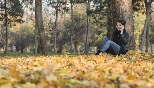Young woman using phone while sitting by tree trunk during autumn
