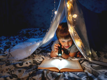 Cute boy reading book with flashlight at night