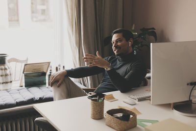 Smiling businessman talking through wireless in-ear headphones while sitting at desk in office