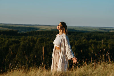 Happy young woman meditating and catching sunlight outdoors at sunset with scenic landscape