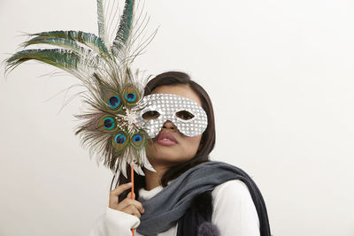 Portrait of young woman wearing eye mask holding peacock feathers against white background