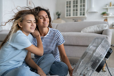 Optimistic relaxed mother and daughter enjoy wind coming from fan sits on floor in living room