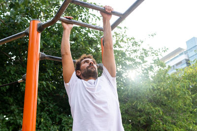 From below hispanic male athlete exercising on monkey bars against lush trees during fitness workout on sports ground in park