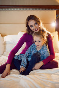 Mother in purple clothes with a sad dissatisfied daughter in a dress is sitting on a bed in a hotel