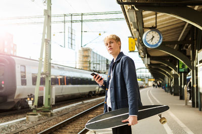 Young man holding mobile phone and skateboard while waiting on railroad station platform