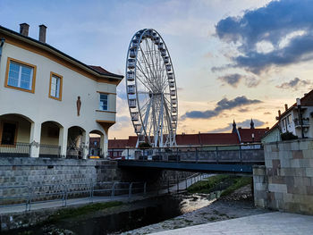 Ferris wheel by river and buildings against sky