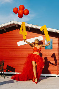 Trendy black female with decorative numbers and red balloons standing on walkway against construction during birthday party in sunlight