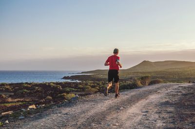 Rear view of man running on dirt road on coastline against sky