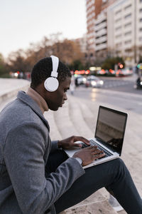 Young man wearing headphones working on laptop while sitting in city